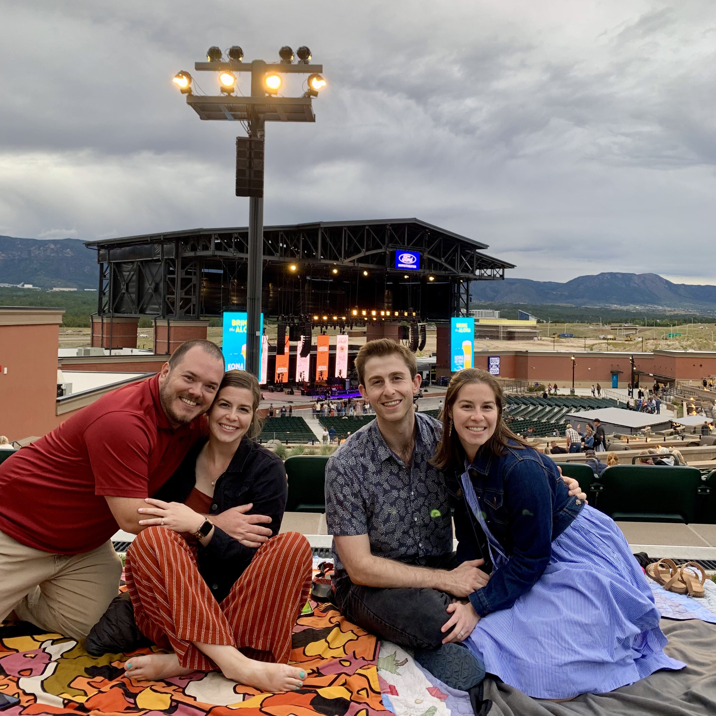 Two couples sitting on a blanket in front of an outdoor stage with mountains in the background – turn your favorite memories into custom photo magnets by Spellbound Magnet Co.