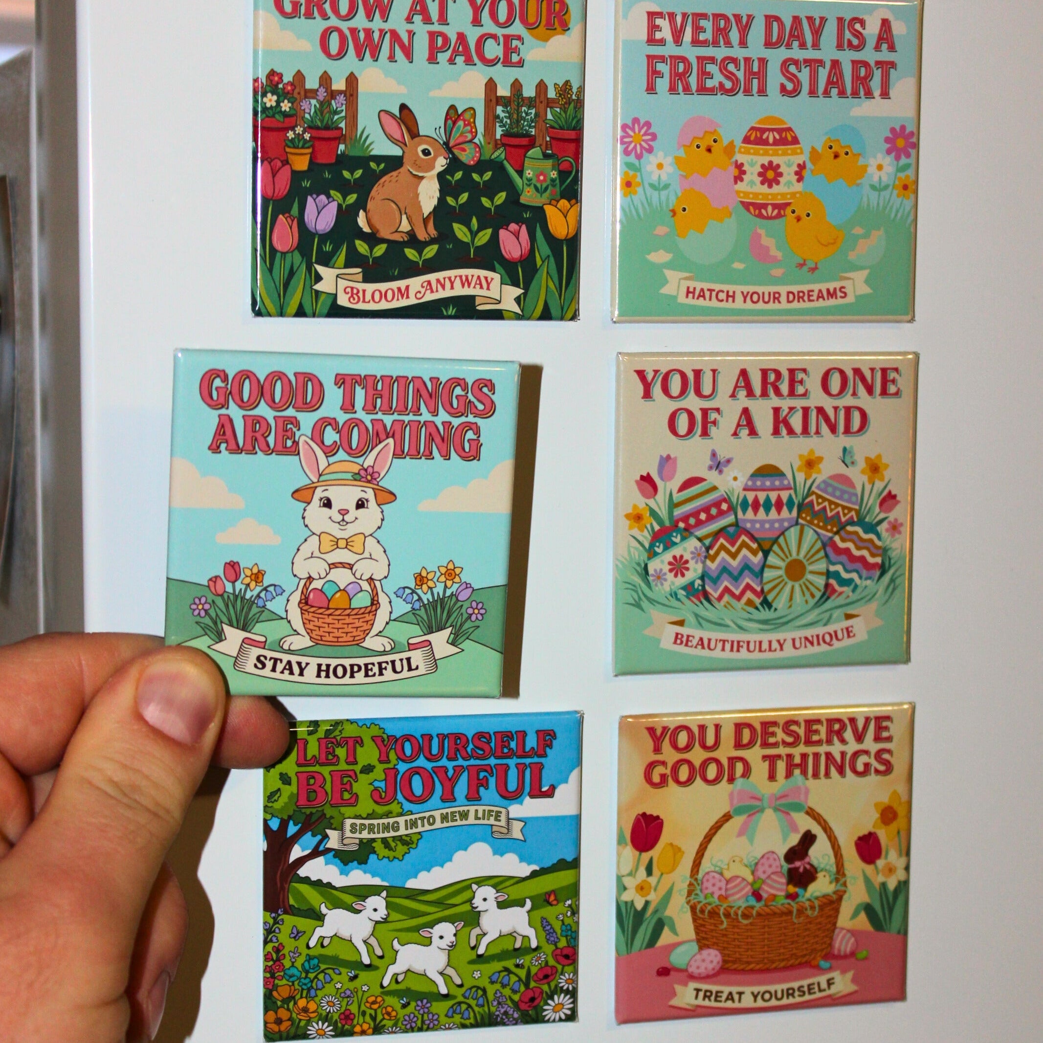 Hand holding the Good Things Are Coming / Stay Hopeful magnet in front of a white refrigerator displaying all six Spellbound Magnet Co. Easter 2026 Magnet Pack designs, including Grow at Your Own Pace, Every Day Is a Fresh Start, You Are One of a Kind, Let Yourself Be Joyful, and You Deserve Good Things.