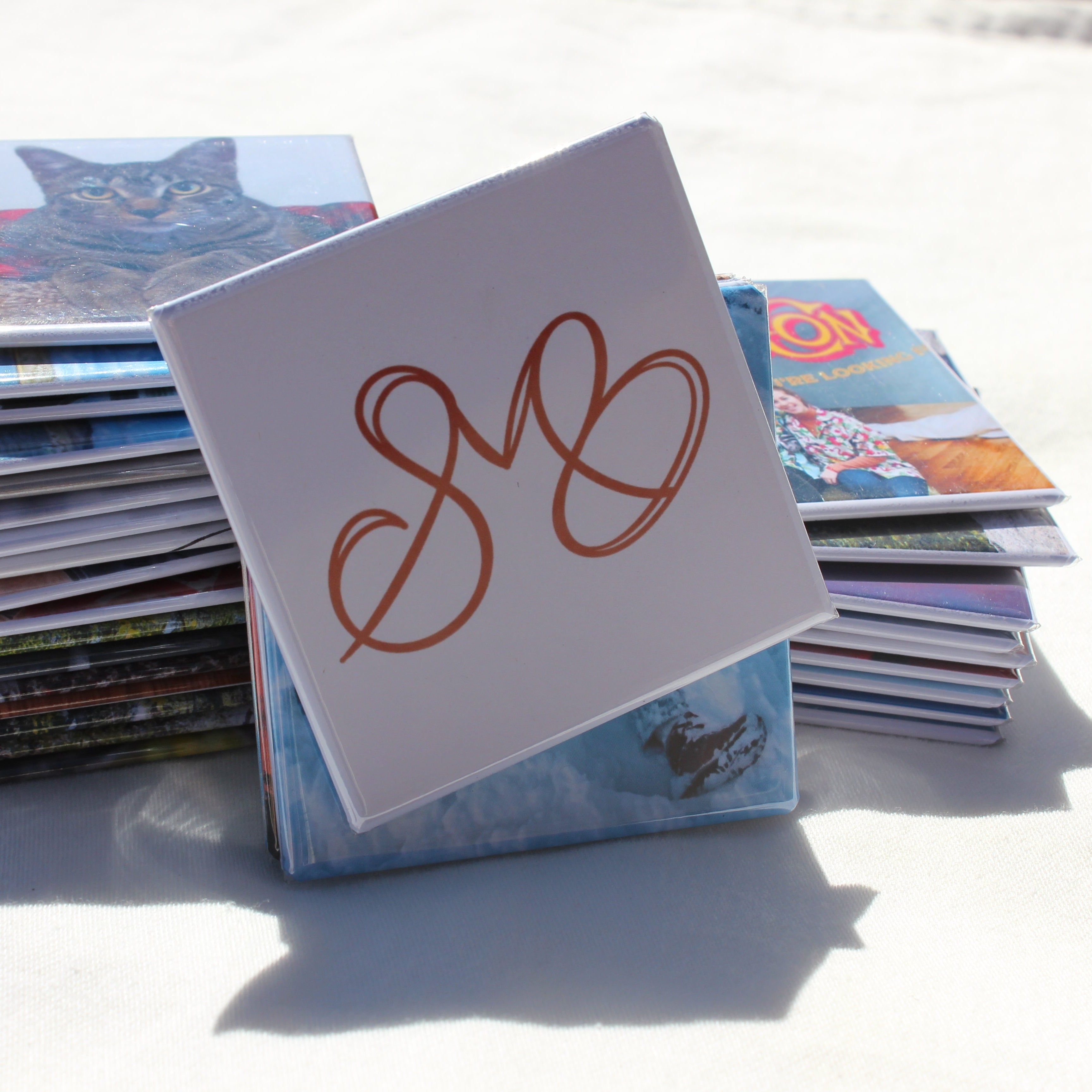 Spellbound Magnet Co. logo magnet leaning against a large stack of custom photo magnets – 2.5 inch square photo magnets handmade by Spellbound Magnet Co.